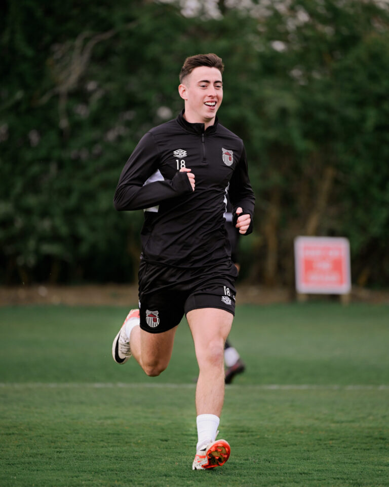 This image shows a person wearing a black athletic training top and shorts with the crest of Grimsby Town F.C. while running on a turf pitch.